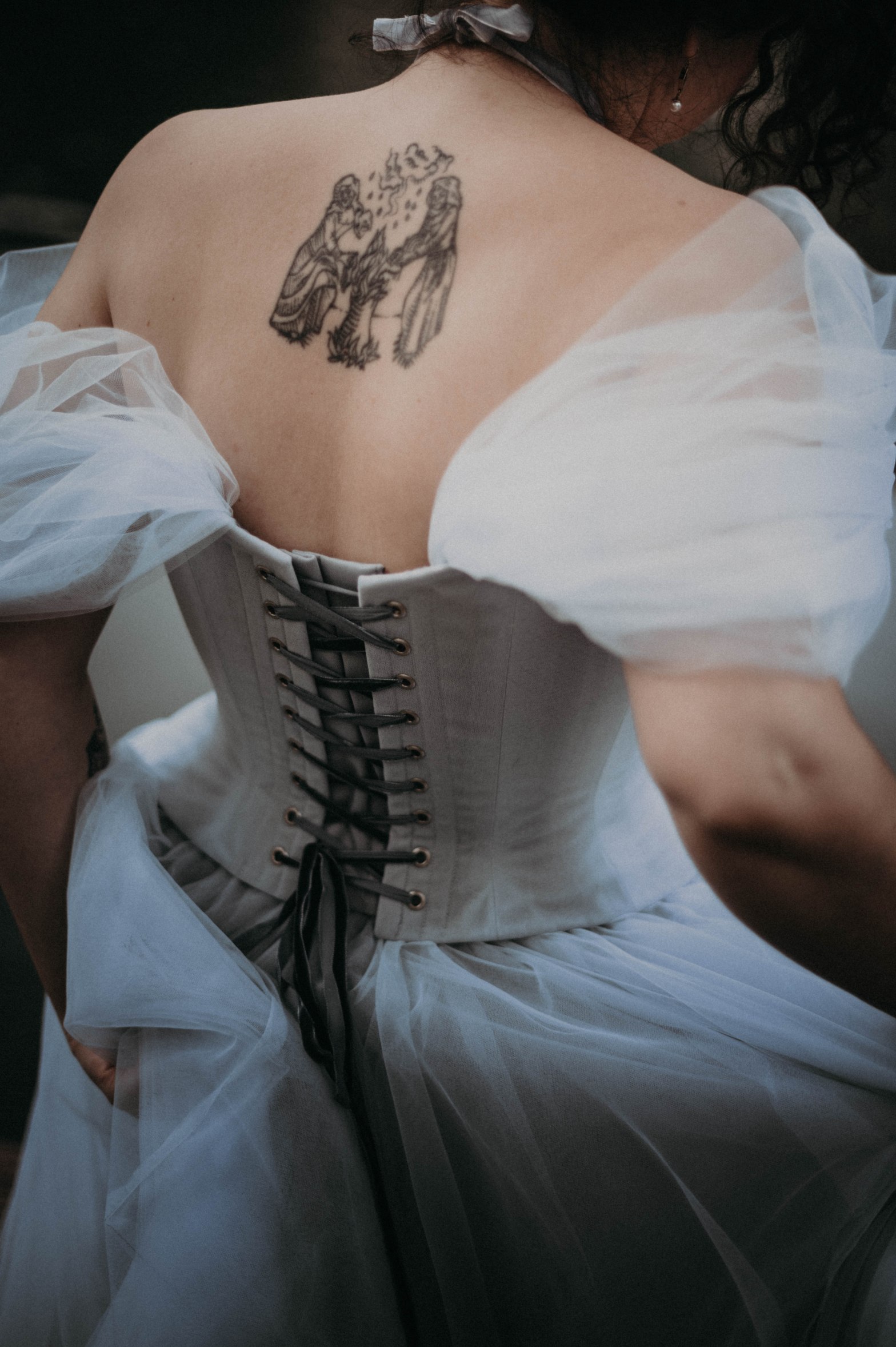Photo d'une robe de mariée corsetée de dos. La robe est en tulle bleu gris et on voit un tatouage dans le dos de la modèle ainsi que le laçage du corset.
