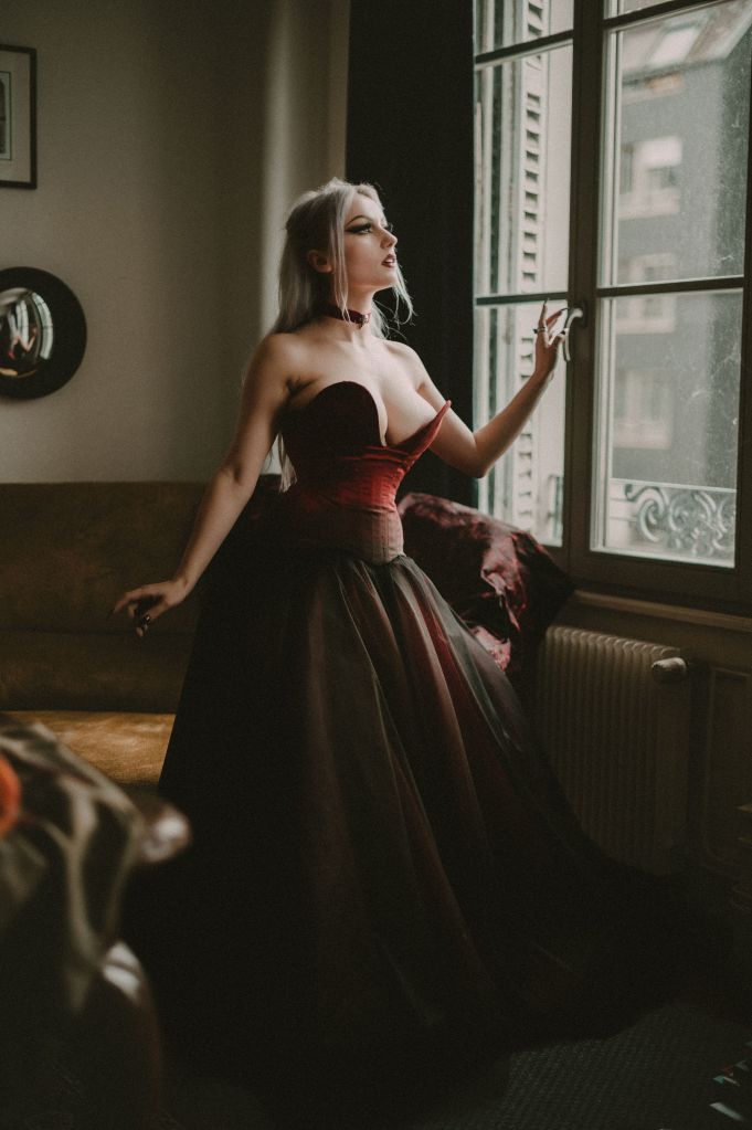 Une modèle dans un style gothique avec les cheveux argentés raides pose devant la fenêtre d'un appartement dans une ambiance victorienne. Elle porte un corset en dupion de soie sauvage dans un dégradé allant du rouge au noir. Elle porte une jupe vaporeuse en tulle rouge et noir.
