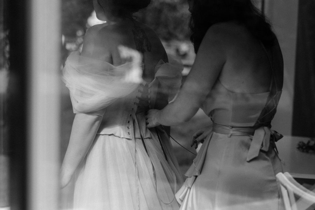 Photo en noir et blanc, une demoiselle d'honneur lace la robe corsetée de la future mariée.